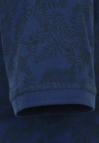 Marineblå stof med en struktureret vævning og et mørkegrønt bladmønster. Ærmehullet er ribbet, hvilket tilføjer struktur til designet.