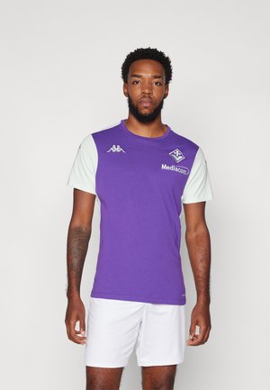 Uomo in piedi con una maglia sportiva viola e bianca con il logo Mediacom e pantaloncini bianchi su uno sfondo semplice.