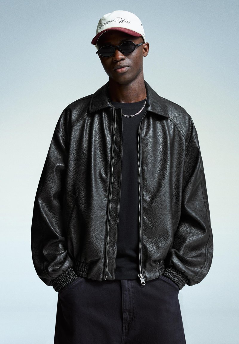 Giacca bomber in pelle nera con zip frontale e polsini elasticizzati, abbinata a una maglietta nera e pantaloncini scuri, accessoriata con occhiali da sole e un cappellino.