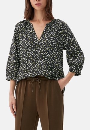 Žena nosící černou, bílou a olivově květovanou halenku s výstřihem do V, 3/4 nafouklými rukávy a hnědé kalhoty se šňůrkou, ruka v kapse, viditelný trup.