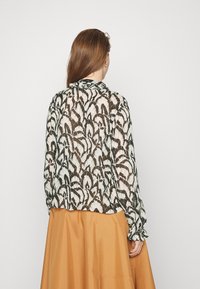 Blusa in pizzo con un motivo floreale in nero e crema, maniche lunghe e polsini arricciati, abbinata a una gonna marrone uniforme.