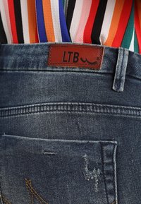 Jeansy dżinsowe w wyblakłym niebieskim kolorze, z brązową skórzaną łatą oznaczoną napisem „LTB” oraz tylną kieszenią z delikatnym przeszyciem.