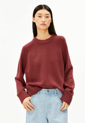 Jonge vrouw met lang donker haar draagt een losse bordeauxrode trui en lichtblauwe high-waisted jeans, staand met handen in de zakken.
