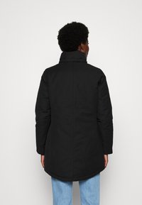 Cappotto nero di lunghezza media con colletto alto, texture liscia e maniche lunghe. Presenta un orlo curvo e un design minimalista.