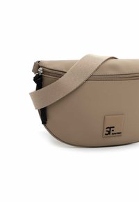 Beige crossbodytas met een gladde, matte textuur, ritssluiting en een platte band. Heeft een logopatch aan de voorkant en zwarte metalen accenten.