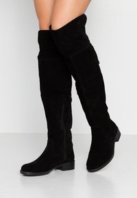 Botas negras de ante por encima de la rodilla, con un diseño elegante, punta afilada y cremallera lateral. Tacón plano para mayor estabilidad.