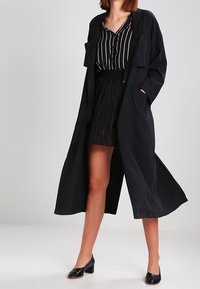 Long manteau trench noir à coupe décontractée, associé à une chemise rayée noir et blanc et une jupe noire plissée. Chaussures noires à talons.
