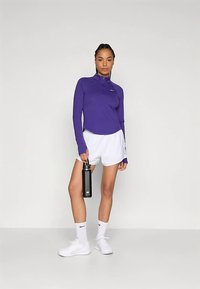 Lilla langermet sportstopp med halvlukking, kombinert med hvite shorts. Modellen holder en svart vannflaske, og har på seg hvite sokker og sko.