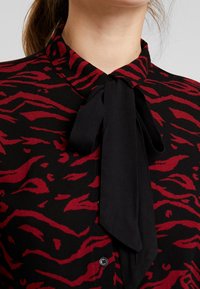 Blusa nera con un motivo a strisce di tigre rosse, caratterizzata da un colletto alto e un lungo fiocco nero da annodare al collo. Tessuto liscio, chiusura a bottoni.