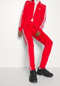 Röd träningsoverall med vita ränder, bestående av en zip-jacka och matchande byxor. Bärs med svarta sportskor. Slät textur, atletisk design.