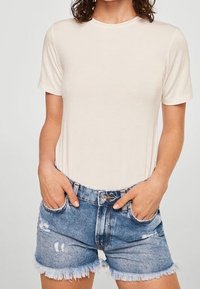Camiseta de manga corta beige en tejido suave, combinada con shorts de denim azul desgastado con dobladillos deshilachados y bolsillos frontales.
