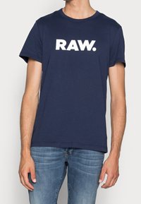 Náměsíčná modrá bavlněná tričko s krátkým rukávem, se výrazným bílým logem "RAW." na přední straně. Hladká textura, standardní střih.