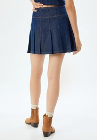 Jupe en denim plissée de couleur bleu foncé, longueur mi-cuisse, avec des coutures visibles. Assortie à des bottes cheville en daim marron et des chaussettes crème.