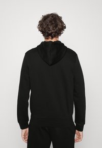 Uomo con capelli ricci che indossa un semplice hoodie nero e pantaloni neri, in piedi di spalle contro uno sfondo grigio chiaro.