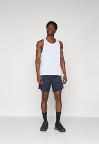 White tank top, dark shorts, black athletic shoes, and black socks. Model has short curly hair and stands against a light gray background.