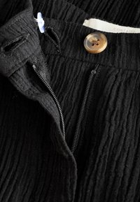 Pantalon en tissu côtelé noir avec un bouton marron, une fermeture éclair partiellement ouverte et un bouton en plastique bleu fixé près de la ceinture.