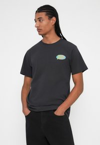Sort bomulds T-shirt med korte ærmer, rund hals og et farverigt grafisk logo på brystet. Modellen har sorte bukser på.