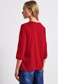 Rotes, langärmeliges Oberteil mit lockerer Passform, Rundhalsausschnitt und gerafften Bündchen, kombiniert mit blauen Denim-Jeans, von hinten aufgenommen.