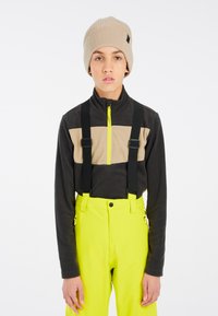Dunkelgrauer Fleece-Oberteil mit beiger Streifen und Reißverschlusskragen; kombiniert mit hellgelben Hosen mit verstellbaren schwarzen Hosenträgern. Beiger Beanie als Akzent.