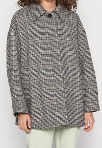 Veste surdimensionnée à motif pied-de-poule noir et blanc, avec un grand col, une fermeture par un bouton unique et une coupe ample. Portée par-dessus un pantalon vert clair.