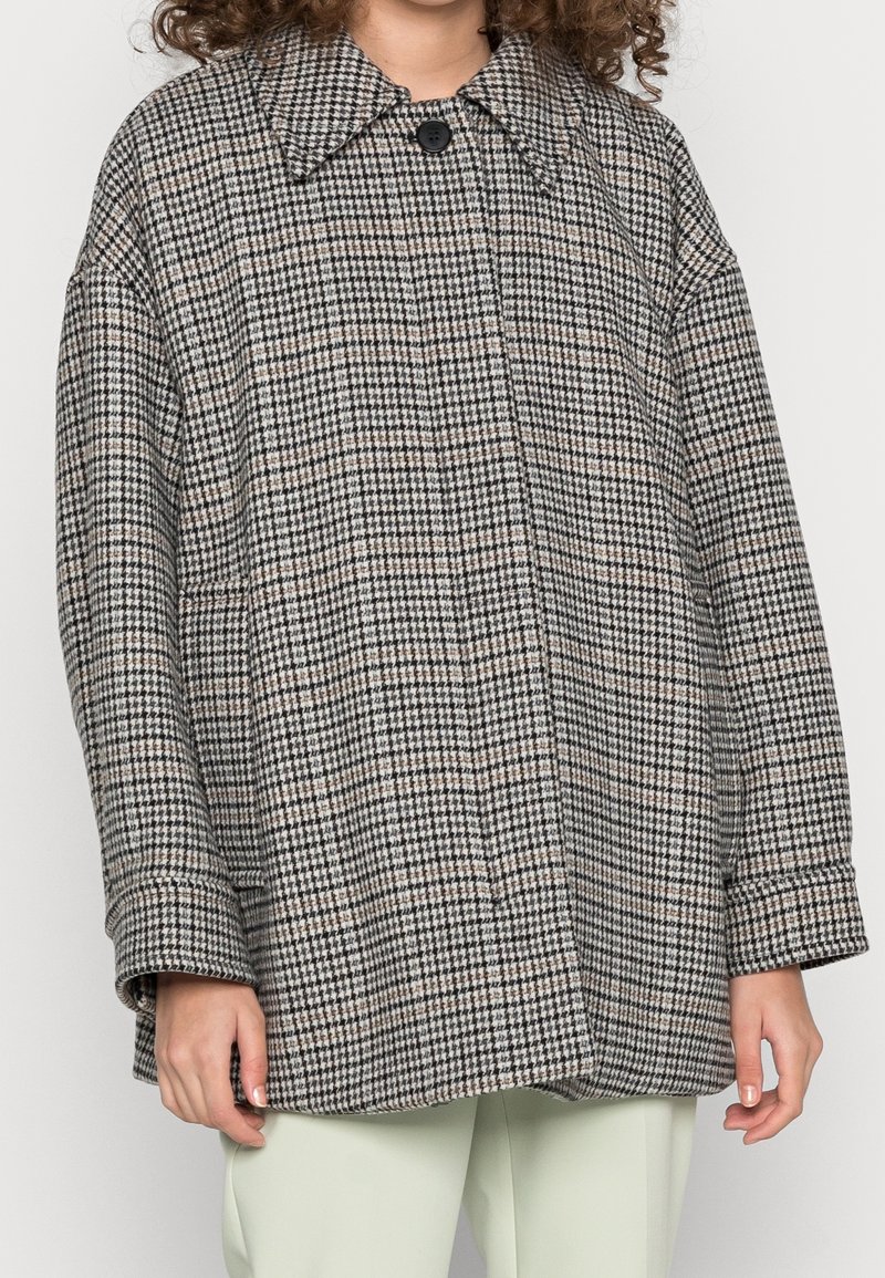 Veste surdimensionnée à motif pied-de-poule noir et blanc, avec un grand col, une fermeture par un bouton unique et une coupe ample. Portée par-dessus un pantalon vert clair.