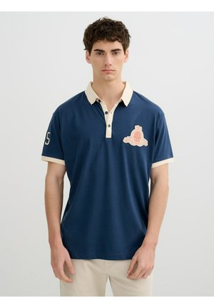Joven con pelo rizado corto lleva una camiseta polo azul marino con cuello y emblema crema, de pie frente a un fondo claro y liso.