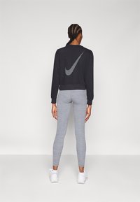 Kobieta z warkoczami stoi tyłem, ubrana w czarną bluzę z dużym białym logo Nike, szare legginsy i białe sneakersy.