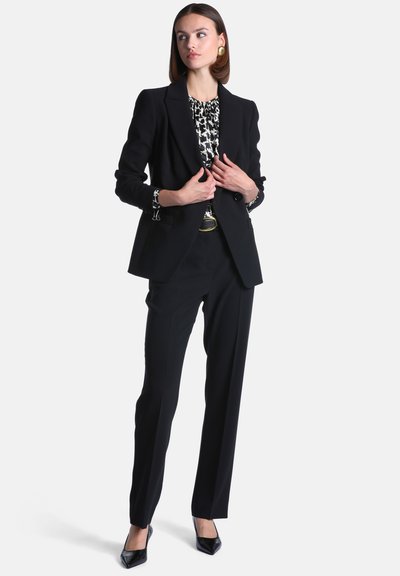 Femme portant un costume noir sur mesure avec un chemisier noir et blanc à motifs, tenant les revers de son blazer, debout devant un fond uni.