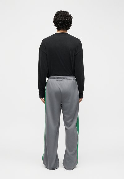 Άνδρας με σγουρά μαλλιά που φοράει μαύρη μπλούζα με μακριά μανίκια και γκρι παντελόνι φαρδιά με πράσινες ρίγες στο πλάι, στέκεται γυρισμένος με την πλάτη σε λευκό φόντο.