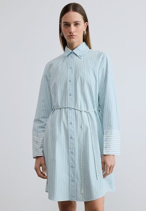 Frau trägt ein hellblaues, vertikal gestreiftes Hemdblusenkleid mit großen Manschetten und einem dünnen Gürtel, der in der Taille gegen einen weißen Hintergrund gebunden ist.