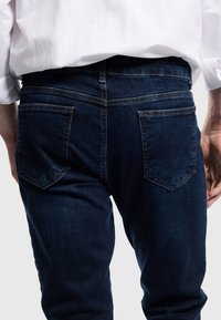 Tummansinisiä farkkuja, joissa on vartalonmyötäinen malli, kaksinkertaiset takataskut ja kontrastiväriset ompelu yksityiskohdat. Kankaan pinnalla on hieman teksturoitu ilme.