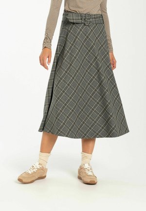 Mujer con zapatillas beige, calcetines crema, una falda midi a cuadros gris con cinturón y una blusa estampada de manga larga, de pie con las piernas separadas.