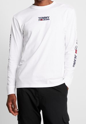 Mężczyzna w białej, długorękawowej koszuli Tommy Jeans z logo na piersi i rękawach, w połączeniu z czarnymi spodniami typu cargo, stojący na jednolitym tle.