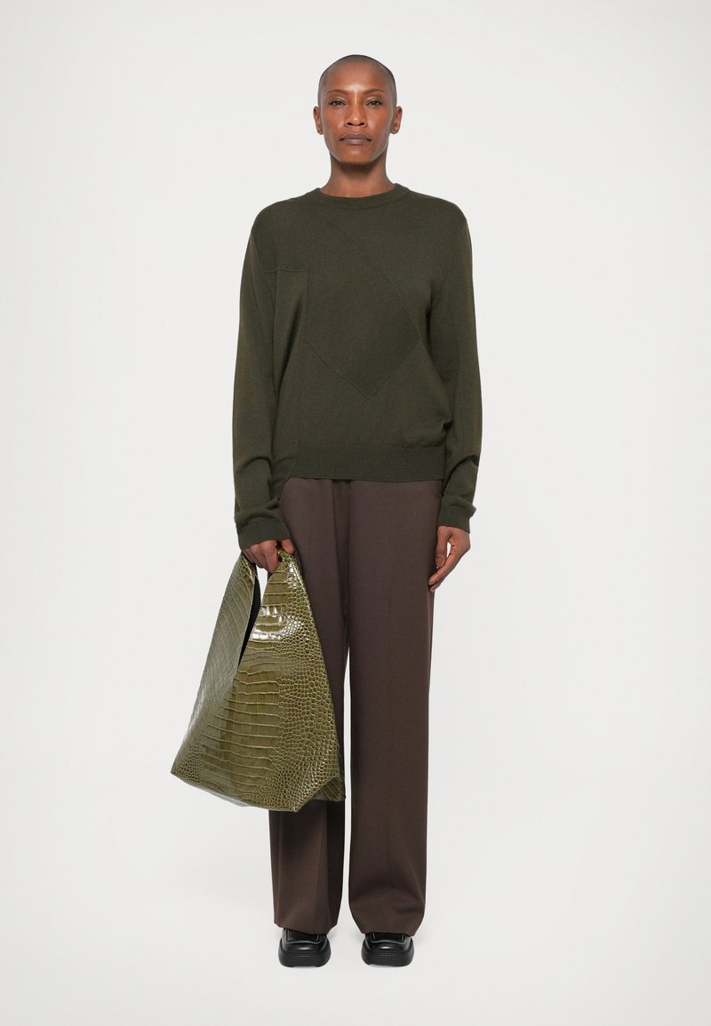 Un pull vert olive avec un motif en diamant texturé, associé à un pantalon large marron. Tenant un sac en croco brillant de couleur vert clair.