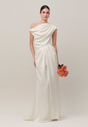 Frau mit dunklen Haaren in einem weißen Satin-Ein-Schulter-Kleid, die einen kleinen Strauß orangefarbener Rosen vor beigem Hintergrund hält.