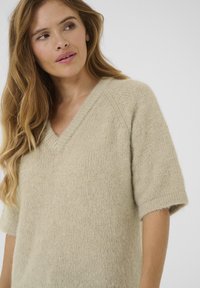Pull en maille beige clair avec un décolleté en V, des manches courtes et une surface douce et texturée. Présente une coupe décontractée et des coutures diagonales subtiles.