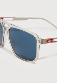 Selge raami prillid siniste läätsede ja metallist templitega, millel on punane HUGO logo valgel taustal.