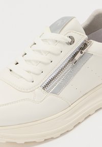 Fehér sneakers, bőr anyagból készült, kerek orral, fűzős záródással, ezüst cipzár díszítéssel és logóval. Texturált talp a jobb tapadás érdekében.