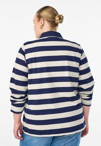 Majica s dolgimi rokavi z navpičnimi črtami v temno modri in kremni barvi. Ima ovratnik, mehko tkanino in sproščen kroj z zavihanimi rokavi.