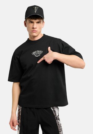 Junger Mann mit schwarzer Kappe und schwarzem Oversize-T-Shirt, der auf ein weißes Logo auf der Brust zeigt, steht vor einem einfarbigen Hintergrund.