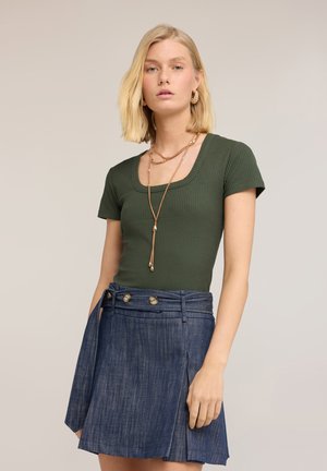 Mujer rubia con top de manga corta acanalado en color verde oliva, collares dorados en capas y falda azul de talle alto con botones sobre fondo liso.