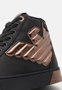 Černá teniska s texturovaným svrškem. Na boku ji zdobí výrazný bronzový kovový motiv křídla a na patě bronzový detail.