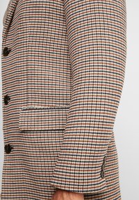 Acercamiento a la manga de una chaqueta de cuadros marrón, marino y blanco con botones oscuros y un bolsillo con solapa en el costado.