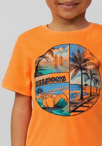 Heldere oranje t-shirt met een ronde afbeelding van strandscènes, palmbomen, surfplanken en de tekst "SUNSET SESSIONS" in vet zwart.