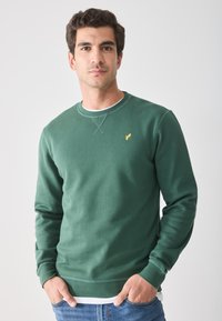 Grüner Baumwollpullover mit Rundhalsausschnitt, mit gelbem Logo auf der Brust, gerippten Bündchen und Saum, getragen über einem weißen Hemd.