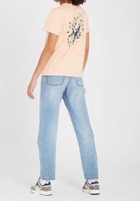 Perzikkleur t-shirt van katoen met een grafische print op de achterkant, gecombineerd met lichtblauwe spijkerbroeken. Model draagt witte en marineblauwe sneakers.