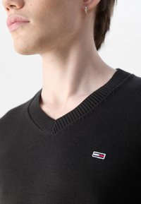 Suéter negro de cuello en V con ribete, que presenta un pequeño logo bordado en rojo, blanco y azul en el lado izquierdo. Tejido suave y texturizado.