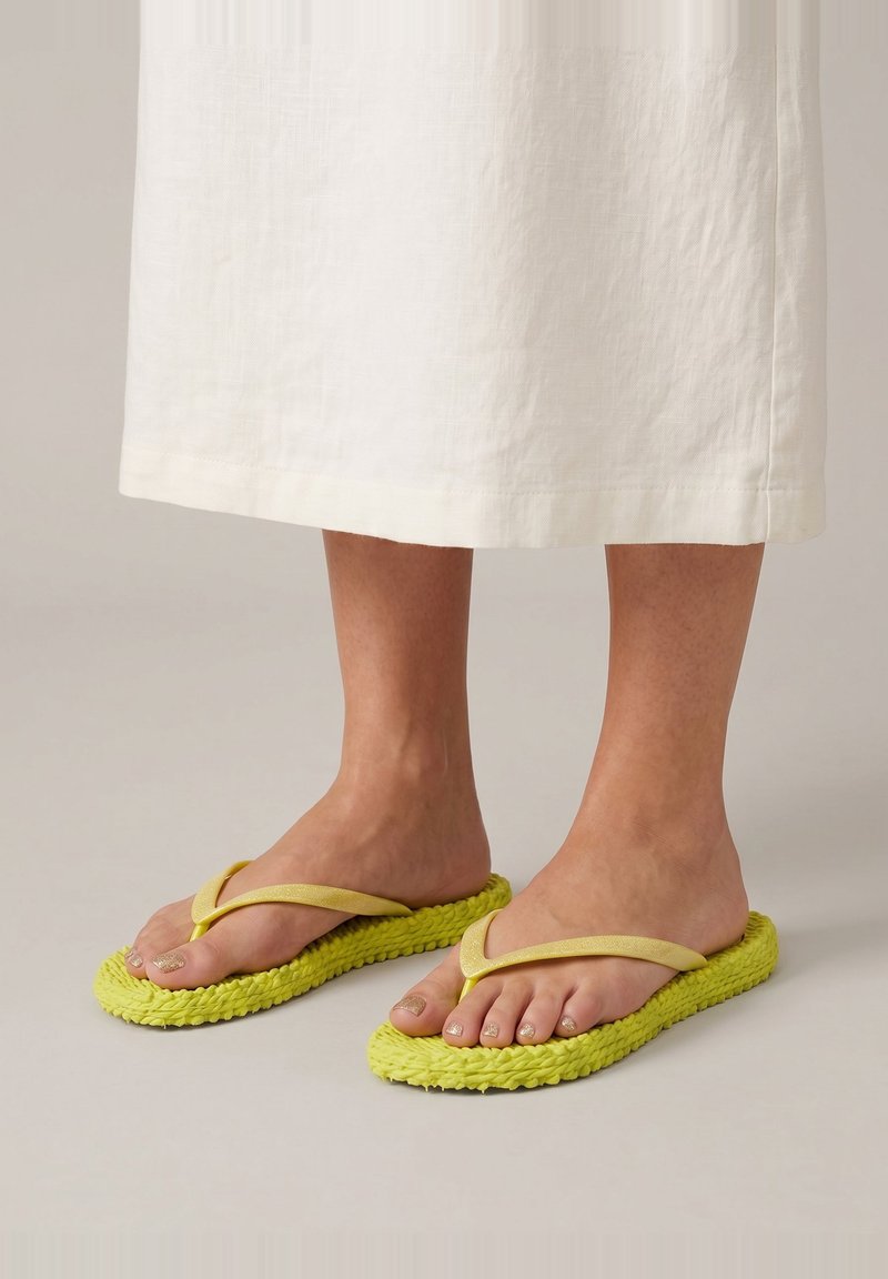 Pieds portant des sandales tongs jaunes avec des semelles tressées et des brides jaune clair, associées à une jupe blanche longueur cheville.