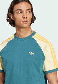 Teal and yellow raglan t-shirt with three yellow stripes on sleeves. Features a small, gold Adidas logo on the chest. Cotton material.