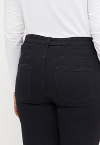 Jeans de denim negro con dos bolsillos traseros, cintura alta y una textura suave. Costuras visibles alrededor de los bolsillos y la cinturilla.